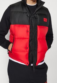 Homme portant une doudoune noire et rouge par-dessus un sweat-shirt noir, les mains dans les poches de la doudoune, avec un patch logo rouge "HUGO" sur la poitrine.
