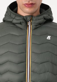 Homme portant une veste matelassée olive avec capuche, fermeture éclair rayée et logo K-Way sur la poitrine et la tirette.