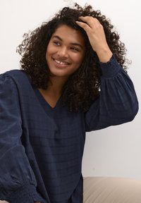 Blusa azul marino de textura, con escote en V y mangas voluminosas y fruncidas. Tela suave con un sutil patrón de cuadros; corte relajado.
