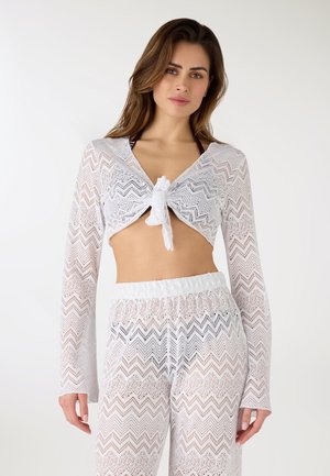 Donna che indossa un top corto a maniche lunghe in pizzo bianco semitrasparente annodato sul davanti e pantaloni in pizzo abbinati a vita alta, su sfondo uniforme.