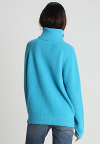 Pull turquoise oversize avec une texture côtelée, un col montant et des épaules tombantes, associé à un jean en denim.