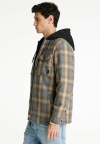 Karierte Flanelljacke in Braun und Grau mit einer schwarzen Kapuze, ausgestattet mit einem Frontreißverschluss, zwei Brusttaschen und einer glatten Textur.