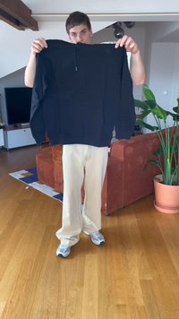 Una persona está de pie en una sala de estar sosteniendo una sudadera negra, lleva pantalones beiges y zapatillas grises, con un sofá rojo y una planta verde cerca.