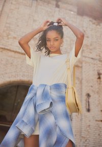 Ljusgul t-shirt, knuten blå rutig skjorta runt midjan, och en liten gul handväska. Långt hår, mot en tegelvägg som bakgrund.