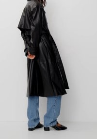Personne portant un long trench noir en cuir, un jean bleu clair et des chaussures à talons bas noires se tenant contre un mur blanc.