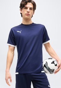 Νεαρός άνδρας με αθλητική ένδυση Puma σε ναυτικό μπλε, κρατώντας μια ασπρόμαυρη μπάλα ποδοσφαίρου μπροστά σε απλό φόντο.