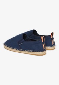Zapatillas slip-on de lona en color navy con suela de yute trenzado, que cuentan con bucles de tiro en naranja y negro en el talón para facilitar su colocación.