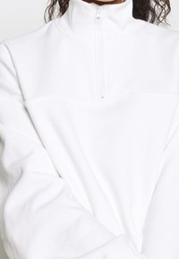 Person klädd i en vit sweatshirt med fjärdedels dragkedja, långa ärmar och hög krage, med hår synligt ovanför kragen.