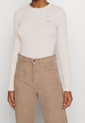 Kobieta ubrana w kremową i jasno-niebieską koszulkę w paski z długim rękawem z logo Levi's oraz beżowe, wysokie spodnie sztruksowe, ręce swobodnie opuszczone wzdłuż ciała.