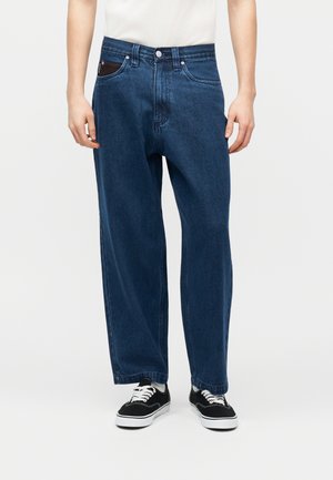 Henkilön alaosa, jossa löysät tummansiniset farkut, valkoinen paitapusero sullottuna housuihin ja mustat tennarit valkoisilla nauhoilla ja pohjilla, yksinkertaisella taustalla.