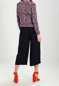 Veste à motifs multicolores avec un design en chevrons rouge, blanc et bleu, associée à un pantalon large noir et des sandales à talons carrés rouges éclatantes.