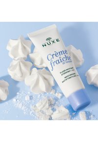 NUXE Crème Fraîche tub på en ljusblå bakgrund, omgiven av vita maränger, som visar en krämfärgad tub med blå kork och etikett.
