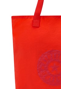 Borsa tote rossa con una finitura lucida, caratterizzata da un design di logo circolare in una tonalità leggermente più scura. Include robuste tracolle in tessuto per il trasporto.