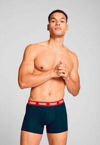 Boxer shorts in dark teal with red elastic waistband featuring "PUMA" logo. Smooth texture, fitted design, mid-thigh length.