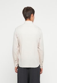 Man wearing a long-sleeve, light beige button-up shirt and dark trousers facing away against a plain white background.