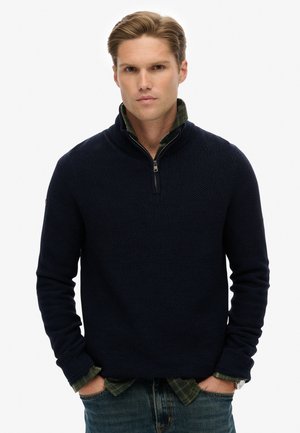 Suéter navy de medio cierre en punto texturizado con cuello y puños acanalados, llevado sobre una camisa de cuadros verde. Combinado con jeans azules.