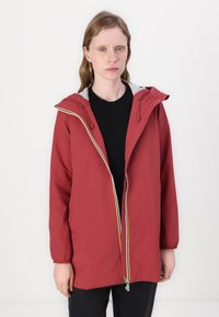 Jachetă impermeabilă roșie cu glugă căptușită cu gri, manșete elastice, două buzunare laterale și un fermoar în contrast. Top negru purtat dedesubt.
