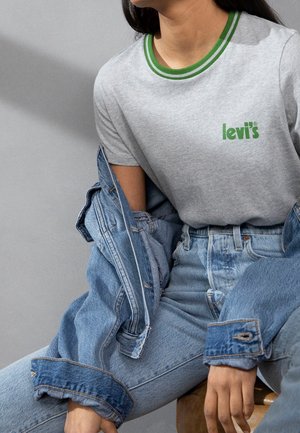 Personne portant un t-shirt gris Levi's avec un bord de col vert, un jean taille haute bleu clair et une veste en jean bleue partiellement portée, assise.