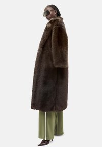 Brun faux fur frakke med høj krave, oversized ærmer og lige snit, båret over løse grønne bukser og spidse brune hæle.