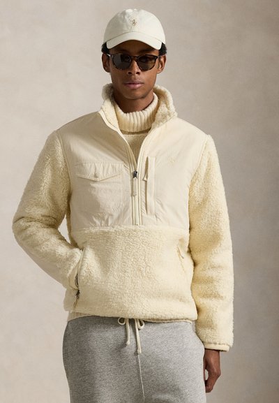 Pull-over en polaire beige clair avec une partie supérieure lisse, fermeture éclair et poches, corps inférieur texturé, porté avec un pull léger et un pantalon gris.