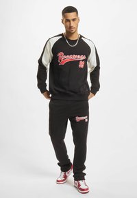 Svart bomullssweatshirt og matchende bukser har rødt "Rocawear 99"-logo, hvite skulderdetaljer og stramme mansjetter. Hvite og røde joggesko fullfører looken.