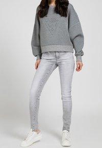 Sudadera gris de gran tamaño con logotipo en relieve, puños y dobladillo acanalados, combinada con jeans skinny gris claro y zapatillas blancas con acentos rosas.