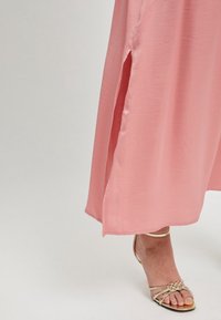 Robe longue et fluide de couleur rose avec une fente sur le côté, fabriquée dans un tissu léger. Associée à des sandales dorées à lanières, mettant en valeur une texture lisse.