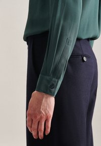 Groene zijden blouse met lange mouwen, knoopdetail bij de manchetten, gecombineerd met donkerblauwe broek met een enkele knoop op de achterzak.