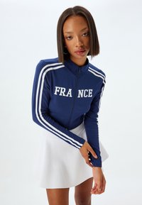 Chaqueta con cremallera en color azul marino con "FRANCIA" en texto blanco, con detalles de rayas blancas a lo largo de las mangas, combinada con una falda acampanada blanca.