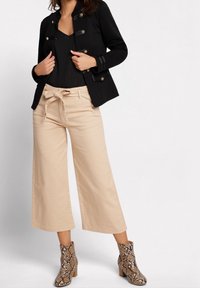 Zwarte getailleerde jas met gouden knopen, gecombineerd met beige wijde broek met een striktaille, en enkelboots met een slangenprintdesign.