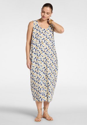 Kvinde iført hvid midi-kjole uden ærmer med blå og gult bladmønster, guldbøjler, armbånd og gyldne snøresandaler, stående.