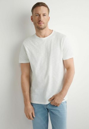 Młody mężczyzna o krótkich jasnobrązowych włosach, ubrany w prostą białą koszulkę z krótkim rękawem i jasnoniebieskie dżinsy, stojący na tle białej ściany.