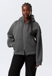 Sweat à capuche gris zippé avec capuche, coupe oversize et poches avant. Poignets et ourlet côtelés, fabriqué dans un tissu doux avec une texture lisse.