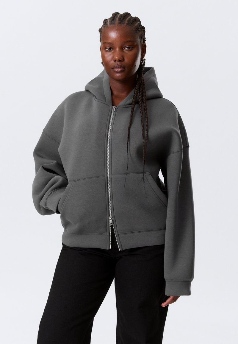 Sweat à capuche gris zippé avec capuche, coupe oversize et poches avant. Poignets et ourlet côtelés, fabriqué dans un tissu doux avec une texture lisse.