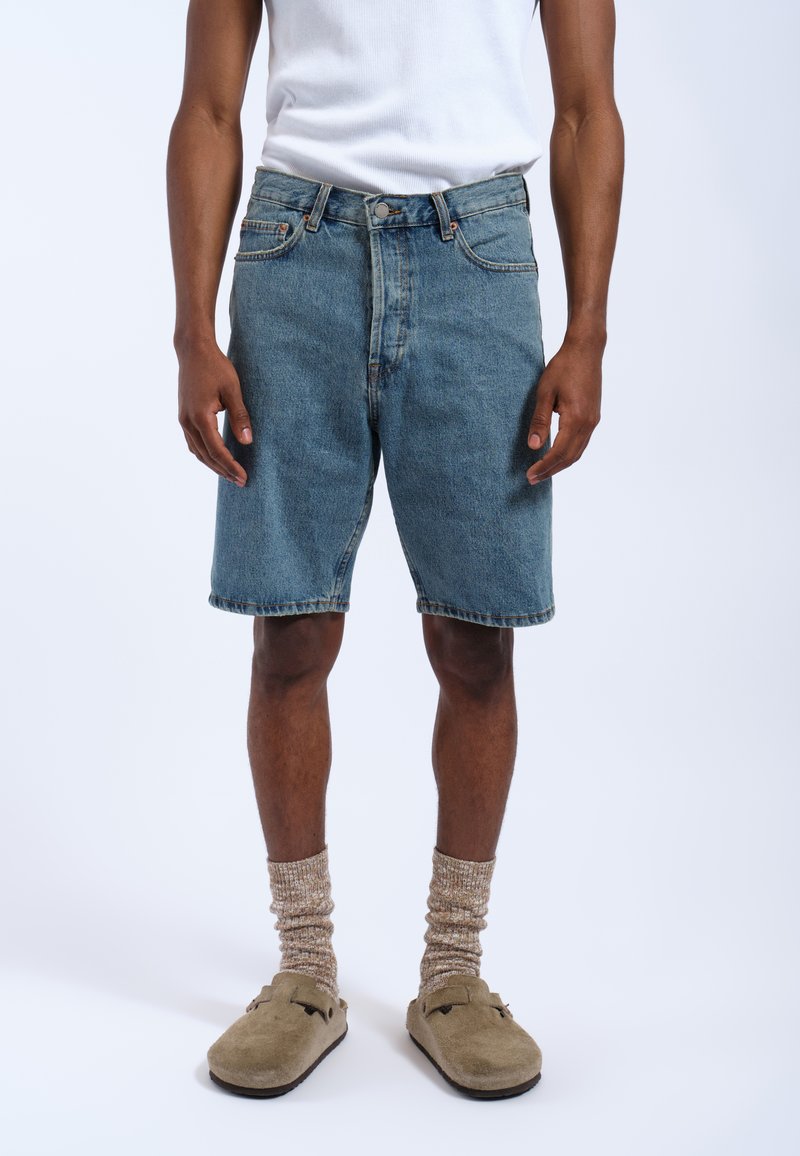 Shorts in denim azzurro chiaro, vestibilità casual, con cinque tasche, passanti per cintura e orlo sfrangiato. Indossati con ciabatte beige e calze a fantasia.