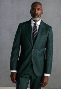 Traje ajustado de color verde oscuro, hecho de un tejido texturizado, con solapa muestrada, cierre de dos botones y bolsillo en el pecho. Camisa de vestir blanca y corbata a rayas.