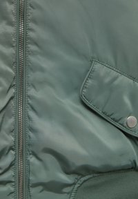 Zelená zateplená bunda s lesklým nylonovým povrchem, sadržující stříbrný zip, boční kapsu na patent a detaily s žebrovanými manžetami.