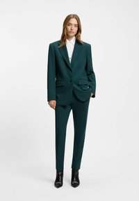 Dunkelgrüner maßgeschneiderter Anzug mit einem strukturierten Blazer, zwei vorderen Taschen, einem Knopfverschluss und passenden schlanken Hosen. Getragen mit einem weißen Hemd.