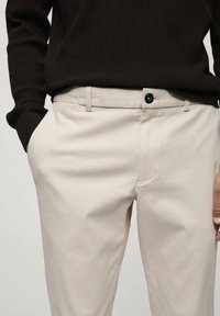 Pantalon beige à devant plat, poches latérales et fermeture par un bouton noir. Le tissu semble lisse avec un léger éclat. Associé à un pull foncé.