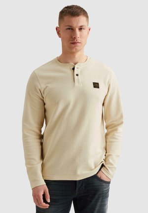 Jonge man die een beige henley-shirt met lange mouwen en twee knopen draagt, samen met een donkere spijkerbroek, staand met één hand in de zak.