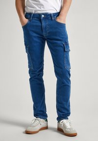 Pantalones cargo de denim azul con un ajuste slim, bolsillos laterales y un ligero estiramiento. Combinados con zapatillas blancas y una camiseta blanca lisa.