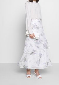 Weiße Bluse mit langen Ärmeln, mehrstufiger Rock mit Blumenmuster in Weiß, Flieder und Grün sowie eine silberne Clutch; kombiniert mit weißen Sandalen mit Absatz.