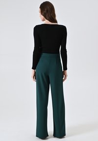 Zwarte geribbelde lange mouwen top gecombineerd met hoge taille, brede groene broeken. Gladde textuur en perfecte pasvorm. Effen achtergrond.