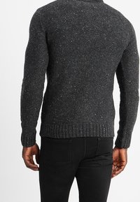 Pull gris foncé texturé avec un motif moucheté, présentant un ourlet et des poignets côtelés, vu de dos. Associé à un jean noir.