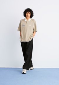 Beige polo shirt with white collar, logo on chest, paired with loose black pants. Model wears neutral sneakers. Simple, relaxed design.