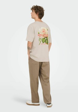 Beige t-shirt met korte mouwen met een grote bloemenprint in kleuren, tekst "Rosa Palida", gecombineerd met losse kaki broek en witte sneakers.