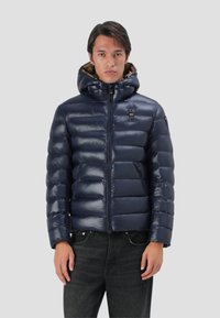 Giubbino trapuntato blu navy con finitura lucida, chiusura con zip, cappuccio attaccato e imbottitura in piuma. Presenta un motivo trapuntato e dettagli con logo.