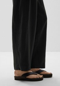 Des sandales en plastique noires avec une large sangle en forme de V, une semelle texturée et une semelle épaisse et rayée. Portées avec un pantalon ample et foncé.