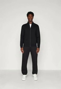 Μαύρο μπουφάν με φερμουάρ και ψηλό γιακά, συνδυασμένο με μαύρους στενούς παντελόνες και λευκά αθλητικά παπούτσια. Λείο ύφασμα, σχεδίαση που κολλάει στο σώμα, ελάχιστες λεπτομέρειες.