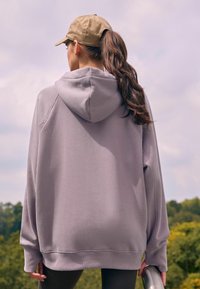 Haut de survêtement oversized violet clair avec une capuche à cordon de serrage et des poignets côtelés, associé à des leggings sombres, tenu par une personne aux longs cheveux.